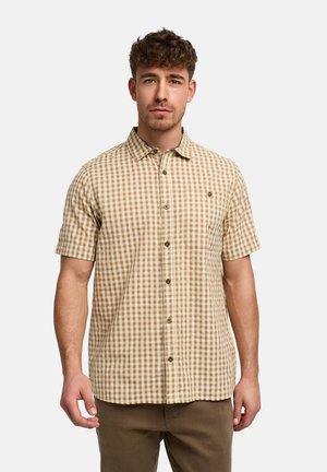 Mężczyzna z krótkimi kręconymi włosami, ubrany w beżowo-białą kraciastą koszulę z krótkim rękawem zapinaną na guziki oraz brązowe spodnie, stoi na białym tle.