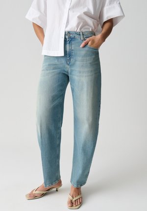 Persona che indossa jeans a vita alta azzurri chiaro, camicia bianca a maniche corte con bottoni e sandali beige con tacco, una mano in tasca, in piedi su pavimento bianco.