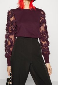 Violetti, pitkähihainen yläosa, jossa on läpikuultavat, röyhelöidyt tummanvioletit verkkomaiset hihat. Yhdistetty korkeavyötäröisiin mustiin housuihin. Musta käsilaukku kädessä.