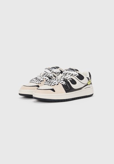 Sneakers in cream and black leather with textured suede accents, featuring thick black-and-white laces and a smiley face logo on the heel.