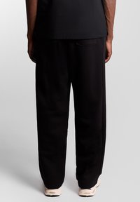 Schwarze Jogginghose mit weiter Passform und elastischem Bund, mit einer Gesäßtasche, kombiniert mit cremefarbenen Sportschuhen. Glatte Textur.