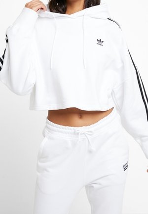 Femme portant un sweat à capuche court Adidas blanc avec des bandes noires et un pantalon de survêtement blanc assorti avec taille à cordon.