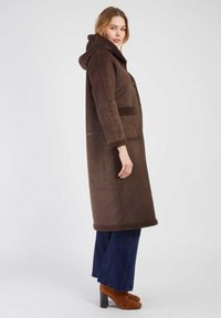 Brązowy zamszowy płaszcz z kapturem, z akcentami z polarowego materiału wzdłuż krawędzi i kieszeni. Długi, prosty krój z widoczną fakturą i detalami przeszyć.