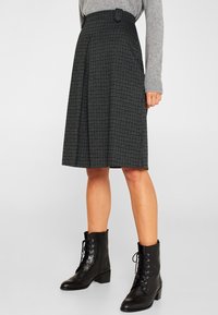 Jupe en plaid noir et gris avec des plis, dotée d'un détail de bouton sur le côté, fabriquée dans un tissu texturé. Associée à des bottines lacées noires.