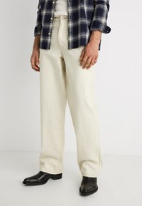 Άνδρας που φοράει εκρού τζιν παντελόνι κανονικής γραμμής, μαύρα δερμάτινα μποτάκια, λευκό πουκάμισο και σκουρό μπλε πουκάμισο με καρό.