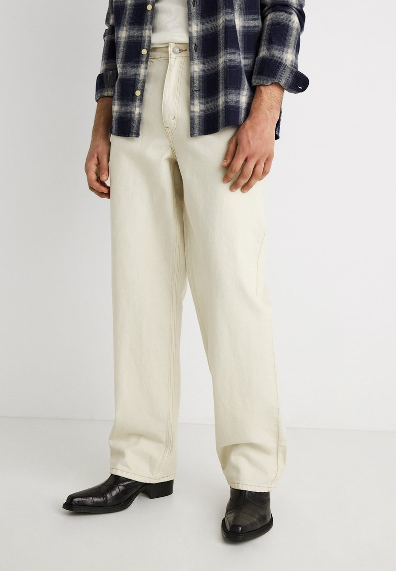 Άνδρας που φοράει εκρού τζιν παντελόνι κανονικής γραμμής, μαύρα δερμάτινα μποτάκια, λευκό πουκάμισο και σκουρό μπλε πουκάμισο με καρό.