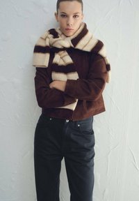 Braune Wildleder-Jacke in Cropped-Optik mit hohem Kragen, kombiniert mit einem gestreiften creme-braunen Schal. Schwarze, hoch taillierte Jeans vervollständigen das Outfit.