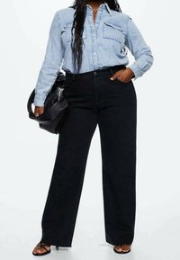 Femme portant une chemise en denim bleu clair rentrée dans un jean noir taille haute à jambes larges, tenant un sac en cuir noir, debout avec une main dans la poche.