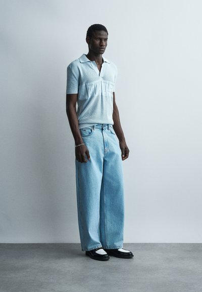 Άνδρας που φοράει ανοιχτό μπλε πόλο μπλουζάκι με κοντά μανίκια, ανοιχτό μπλε τζιν παντελόνι με φαρδιά μπατζάκια και μαύρα-λευκά παπούτσια στέκεται μπροστά σε απλό γκρι φόντο.