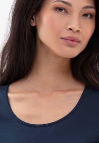 Námořnická modrá blůza s výstřihem ve tvaru lodě vyrobená z měkké tkaniny, s hladkým povrchem a jemným leskem. Výstřih zvýrazňuje oblast klíční kosti.