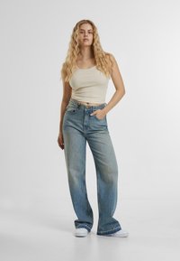 Visoko pašne, svetloblue denim hlače sproščenega kroja, ki vključujejo pet žepek in rahlo zbledenost na stegnih. V kombinaciji s bež topom na naramnice.