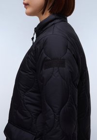 Black quilted jacket with a high collar, featuring a textured pattern and a branded patch on the left arm, made of lightweight material.
