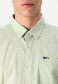 Camisa de mangas curtas verde claro feita de tecido texturizado. Apresenta um colarinho abotoado, bolso frontal e um pequeno logotipo no bolso.