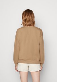 Osoba s vlasy po ramena světle hnědé barvy, oblečená v béžovém svetru a bílých šortkách, otočená zády k jednoduchému světle šedému pozadí.