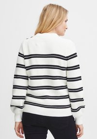 Pull en tricot blanc avec des rayures horizontales noires, doté de poignets et d'un ourlet côtelés. La vue de dos montre un détail de bouton sur l'épaule.