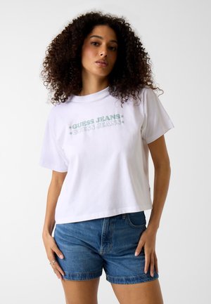 Kobieta o kręconych włosach, ubrana w białą koszulkę "Guess Jeans" i niebieskie denimowe szorty, stojąca na tle jednolitego białego tła.