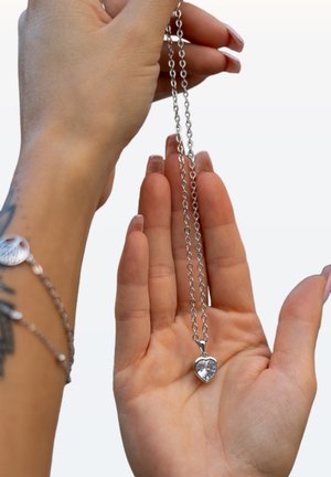 Silberne Kettenhalskette mit einem herzförmigen Anhänger und einem klaren Edelstein. Die Kette hat kleine runde Glieder und wird in einer Hand gehalten.