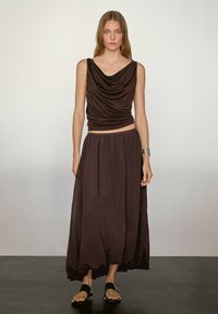 Brauner ärmelloser Oberteil mit gerafftem Ausschnitt kombiniert mit einem langen, fließenden braunen Rock. Das Model trägt schwarze Sandalen und ein silbernes Armband.