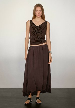 Brauner ärmelloser Oberteil mit gerafftem Ausschnitt kombiniert mit einem langen, fließenden braunen Rock. Das Model trägt schwarze Sandalen und ein silbernes Armband.