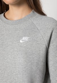 Kvinna som bär en ljusgrå Nike-tröja med vitt broderat märke på bröstet, med detaljer av hals och axlar synliga.