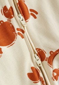Zippered cream garment with orange circular and bunny patterns; soft texture; prominent off-white zipper detail.