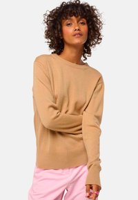 Beiger Pullover aus weichem Strickstoff, runder Ausschnitt, gerippte Saum- und Ärmeldetails, kombiniert mit hellrosa Hosen.