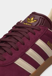 Közeli kép egy bordó Adidas Gazelle sportcipőről, krémszínű csíkokkal, bordó fűzőkkel, arany logóval és egy gumitalppal, fehér háttéren.