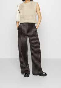 Krémový pletený top bez rukávů s otevřenými vzory, spárovaný s širokými černými pruhovanými kalhotami a robustními černými botami, ruce v kapsách.