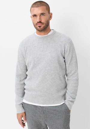 Mann mit kurzem Haar und Bart, der einen hellgrauen Pullover und graue Hosen trägt, steht vor einem schlichten weißen Hintergrund.