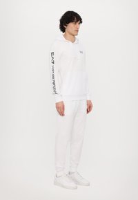 Hvid hættetrøje med kængurulomme og snøre, sort trykt logo på ærmet, parret med matchende hvide joggers og sneaker.