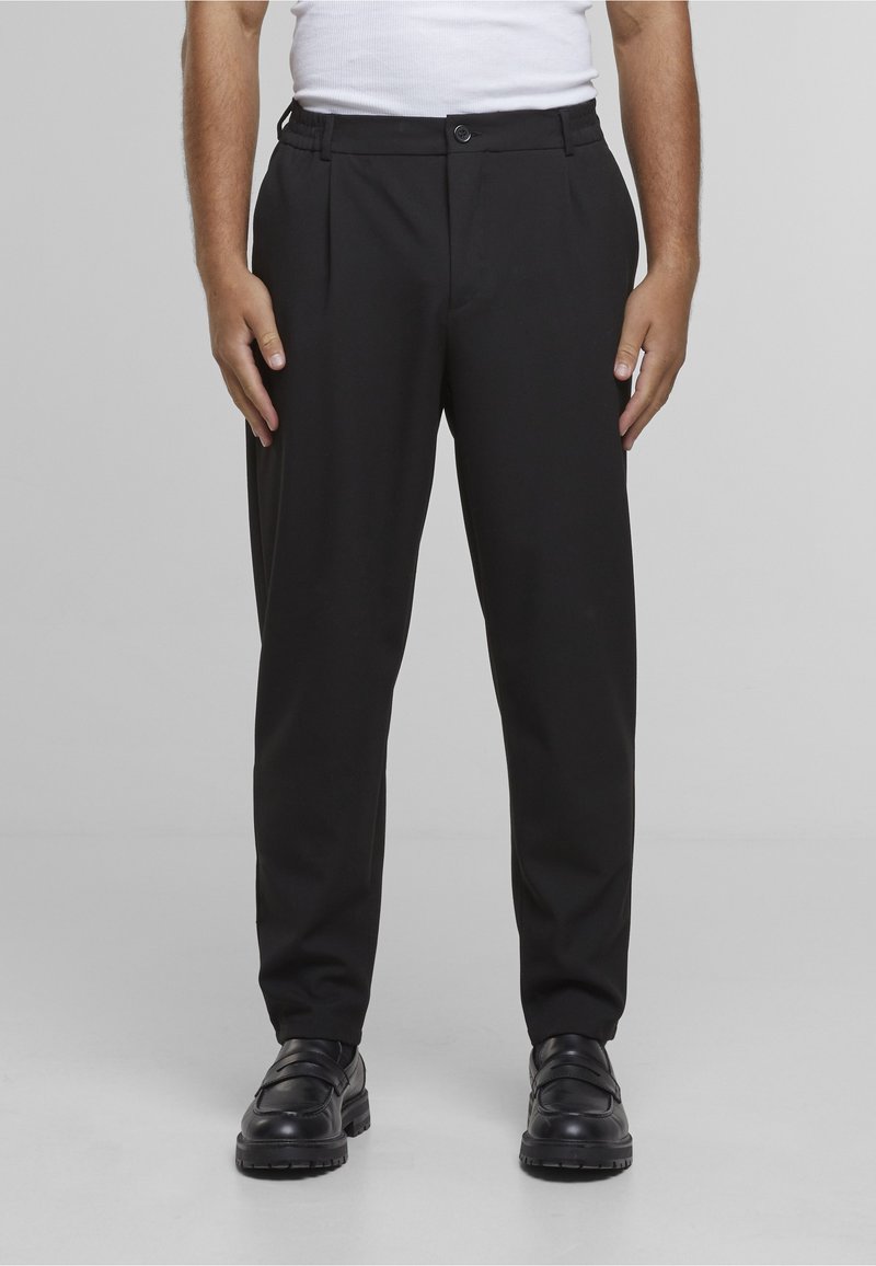 Svarte bukser med avslappet passform, plissert design, elastisk midjebånd, knappelukking og lommer; kombinert med svarte sko for et komplett utseende.