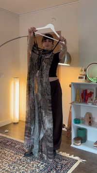 O tânără ținând o rochie lungă, transparentă, cu un model, pe un umeraș alb, într-o cameră confortabilă cu o bibliotecă și o lampă de podea.