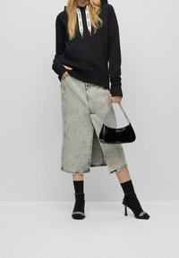 Osoba oblečená v černé mikině s logem na šňůrkách, světle šedé midi sukni s předním rozparkem, černých lodičkách se ponožkami, držící lesklou černou kabelku.