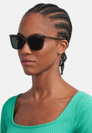 Occhiali da sole neri tartaruga con lenti scure e forma rettangolare. Presentano aste sottili con un accento dorato del logo. Indossati con un top verde a coste.