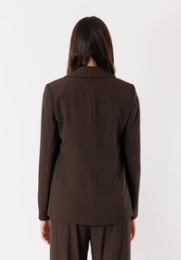 Blazer marrone su misura realizzato in tessuto liscio, con classico risvolto a punta e maniche lunghe, caratterizzato da linee pulite e una silhouette aderente.