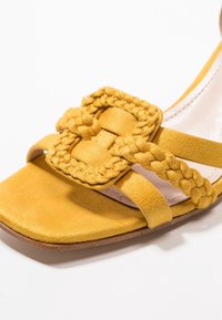 Sandale en daim jaune avec une lanière tissée et une boucle tressée. Le modèle comprend une semelle plate et une doublure intérieure lisse.
