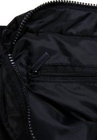 Juodas nailono vidus su užtraukiama kišenė. Savybės: glotni tekstūra ir funkcinė užtrauktuko aparatūra su matinio juodo atsidarymo užtrauktuku.