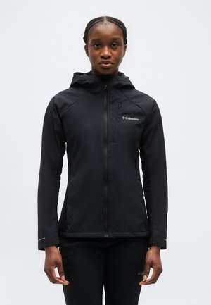 Moteris dėvi juodą „Columbia“ striukę su gobtuvu ir užtrauktuku bei derančias kelnes, stovi priešais paprastą šviesią foną.