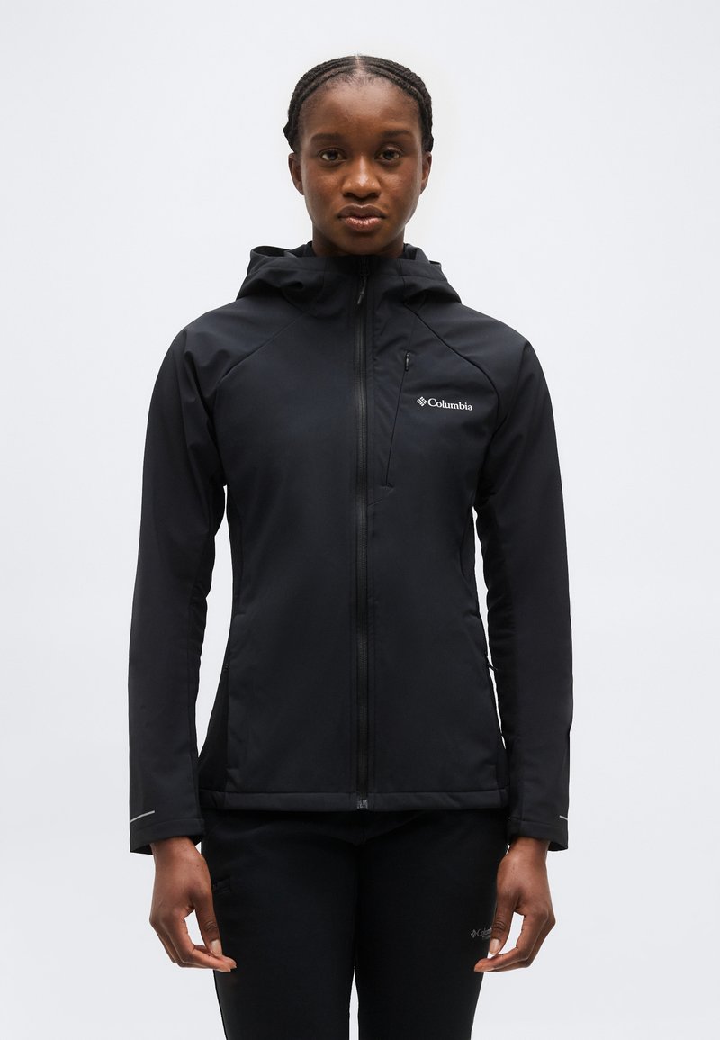 Femme portant une veste à capuche zippée noire Columbia et un pantalon assorti, debout devant un fond clair uni.