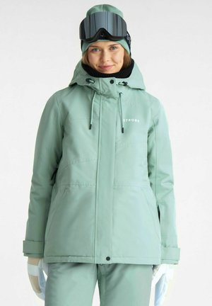 Kvinde iført mintgrøn skijakke, matchende bukser, hue, sort halsvarmer, skibriller på hovedet og hvide handsker mod en ensfarvet baggrund.