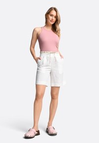 Roze geribbelde tanktop, witte linnenmix shorts met elastische tailleband en zakken, roze klompen met metalen studs, neutrale achtergrond.