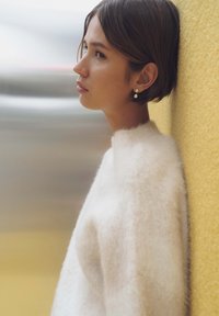 White, fuzzy sweater with a high collar, worn by a person leaning against a yellow wall. Gold stud earrings add detail.