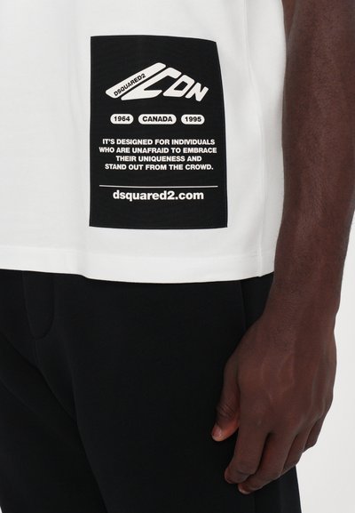 Hvid bomulds t-shirt med et stort sort mærke på siden med tekst, ledsaget af en hånd der rører ved mærket. Sorte bukser er synlige.