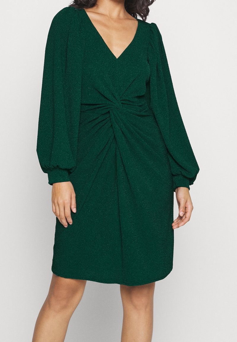 Femme portant une robe longue à manches longues de couleur vert foncé avec un détail torsadé froncé à la taille et un décolleté en V, se tenant contre un fond uni.
