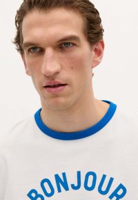 Camiseta blanca de manga corta con cuello de rayo azul y un audaz texto azul "BONJOUR" en un diseño curvado a lo largo del pecho.