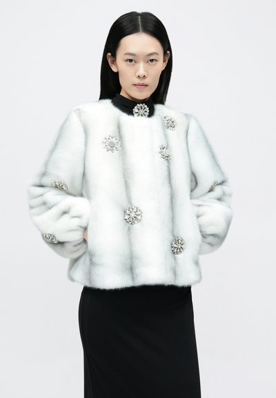 Valge kunstkarusnahast lühike jakk ümarate kaelustega, mis on kaunistatud hõbedaste lillemotiividega ja ühtlase tekstuuriga, sobitatud musta seelikuga.