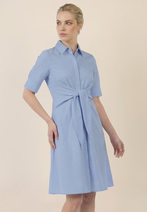 Frau trägt ein hellblaues kurzärmeliges Kleid mit Knoten-Detail vorne, steht und schaut seitlich vor einem schlichten Hintergrund.