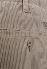 Béžové manšestrové kalhoty s vertikálními žebry, odpovídající knoflík a látková visačka. Textura je měkká a sametová, s viditelnými detaily šití.