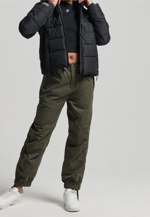 Person iført sort kort pufferjakke, sort kort top, olivengrønne joggerbukser og hvide sneakers mod en ensfarvet baggrund.