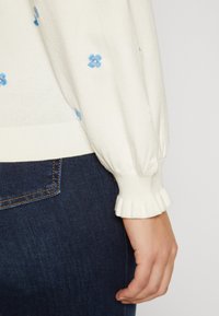 Witte trui mouw met blauwe bloemenborduursel en gerimpelde manchet, rustend nabij de achterzak van donkere spijkerbroek.